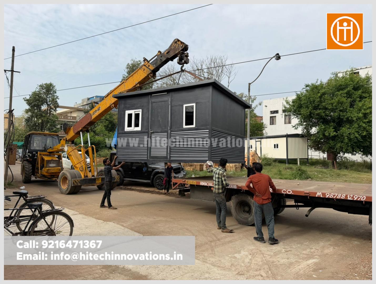 A crane lifting a dark grey prefabricated guard room onto a flatbed truck at the Hitech Innovations workshop.