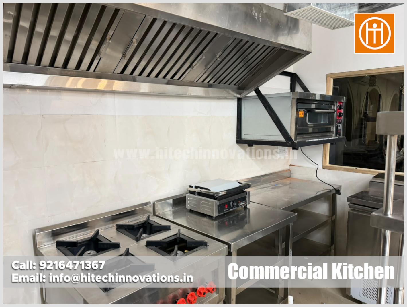 Industrial 4-burner gas range, sandwich griller, wall-mounted commercial oven, and stainless steel exhaust hood in restaurant kitchen