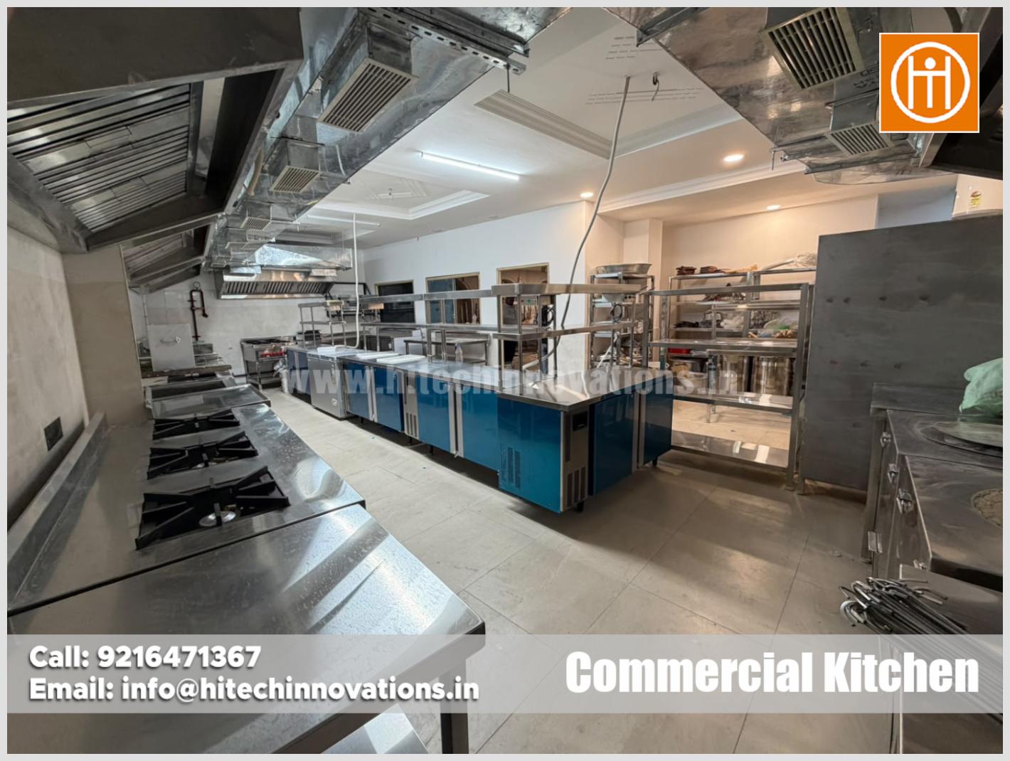 Wide view of commercial kitchen showing stainless steel island work tables, undercounter refrigeration units, and ceiling ducting system